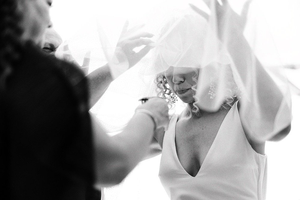 A woman getting ready for a wedding, possibly the bride, with someone assisting her under a veil.