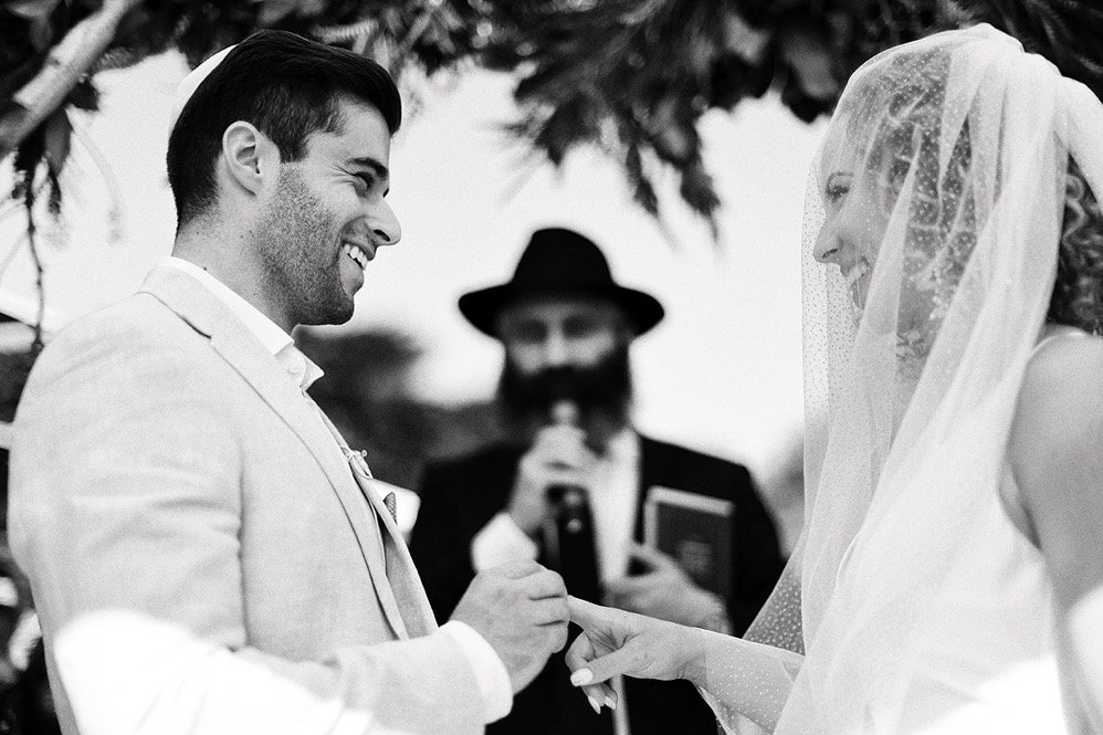 A wedding ceremony with a groom and bride exchanging rings, smiling at each other, under a tree with an officiant in the background.