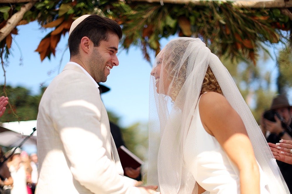 A bride and groom smiling and facing each other during their wedding ceremony outdoors, under a floral arch, with guests visible in the background.