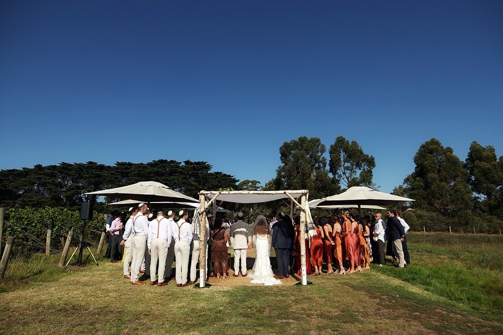 Wedding ceremony outdoors with guests under a white canopy on a grassy field, surrounded by trees and blue sky.