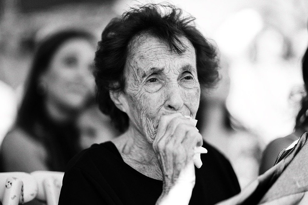 An elderly woman with short dark hair, holding a small piece of paper close to her face, surrounded by other people in the background.
