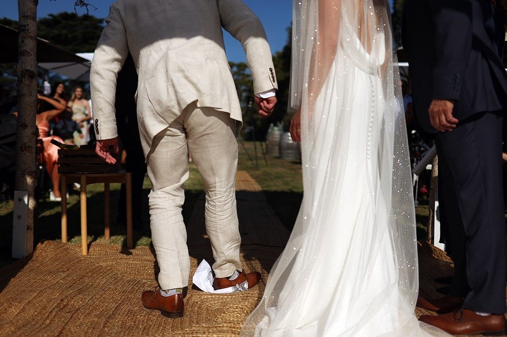 Close-up of a wedding ceremony showing the lower half of the bride and groom, with the bride in a white gown and the groom in a light-colored suit. Guests are seated in the background.