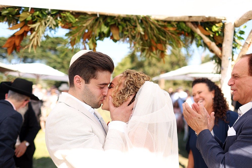 Couple kissing at their outdoor wedding, surrounded by family and friends under decorated archway.