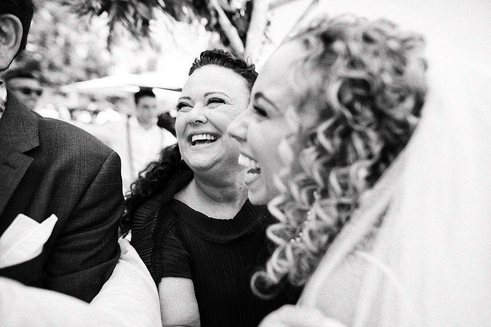 Two women smiling and laughing at an outdoor event, with one woman having curly hair and the other with straight hair and a head covering.