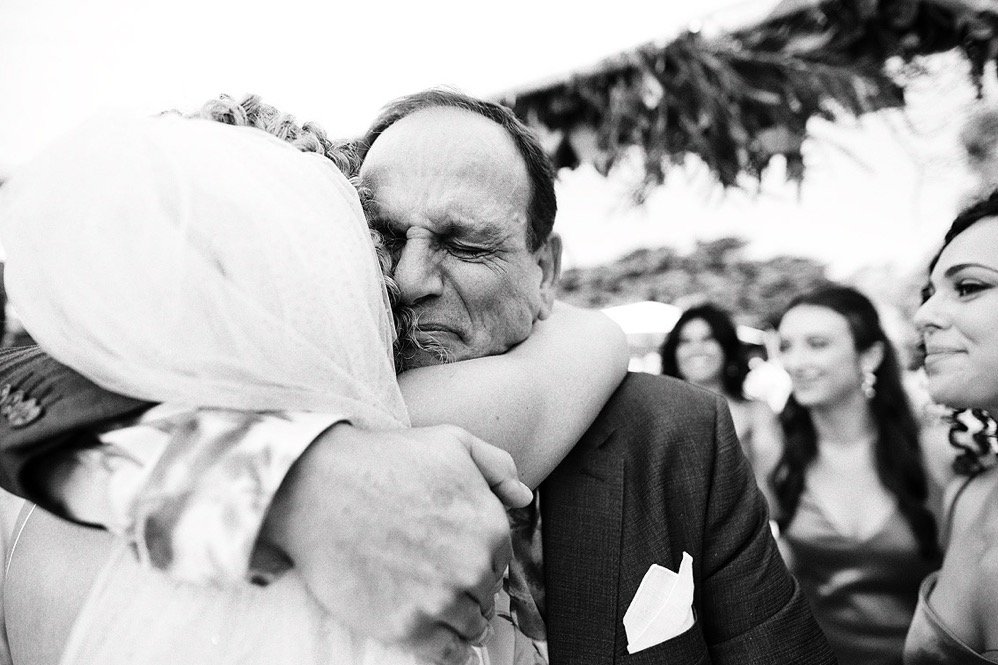 A man and woman hugging, with the man appearing emotional at a gathering or celebration, others smiling in the background.
