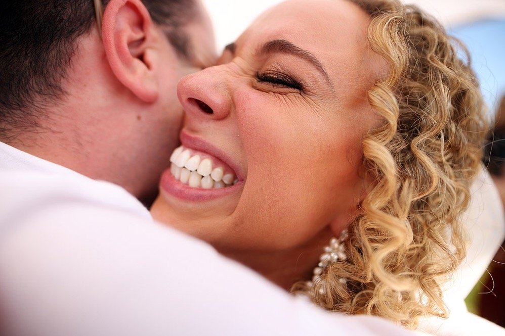 A woman with curly blonde hair and earrings is smiling broadly while hugging a man, whose face is partially visible, showing an intimate moment.