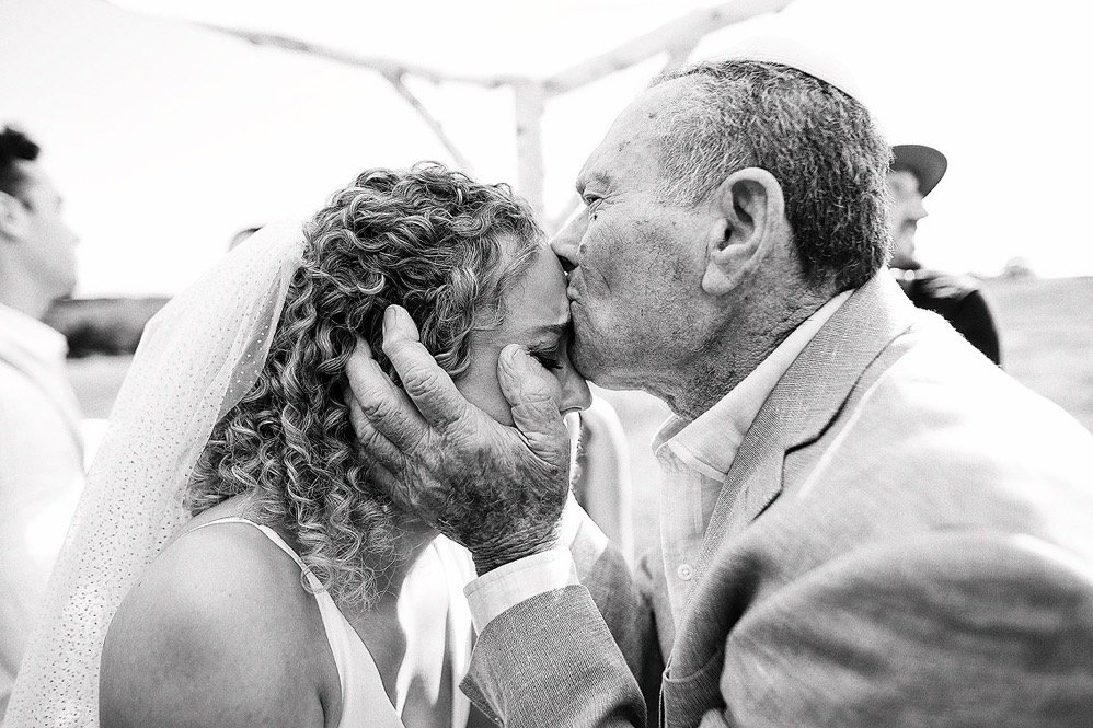 An elderly man kissing a woman on the forehead during a wedding ceremony.