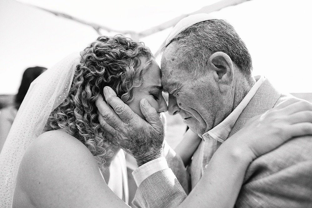 An elderly man and a woman, likely at a wedding, are touching foreheads and holding each other's faces in a tender moment, with their eyes closed in an embrace.