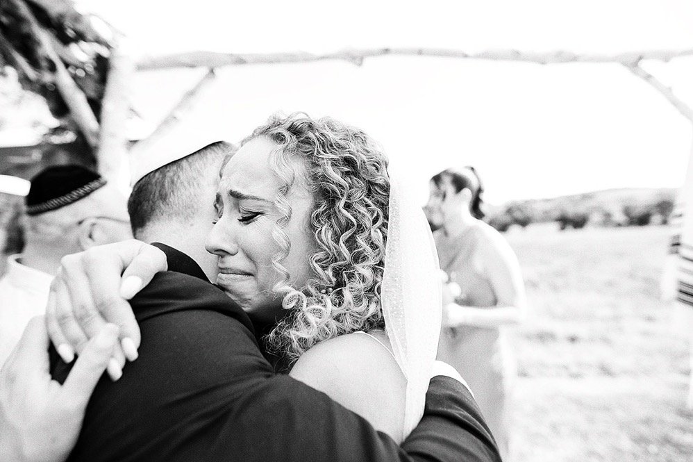A woman with curly hair and a veil embraces a man at a wedding outdoors, with other people in the background.