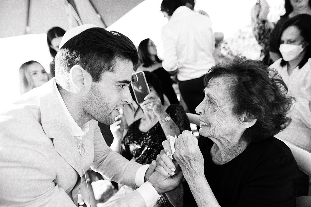 A young man and an elderly woman holding hands and smiling at each other, surrounded by other people in a social setting.