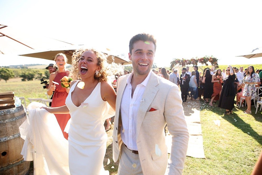 A joyful bride and groom at their outdoor wedding reception, surrounded by guests, with bright sunlight and festive atmosphere.