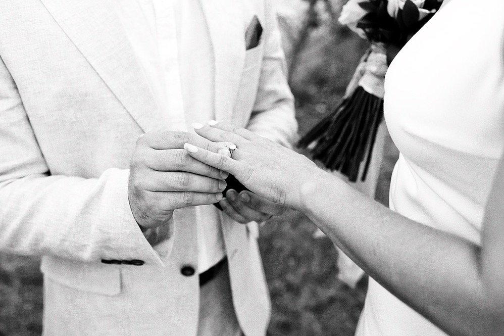 A bride and groom exchange wedding rings outdoors, with the groom holding the bride's hand as he slips the ring onto her finger.