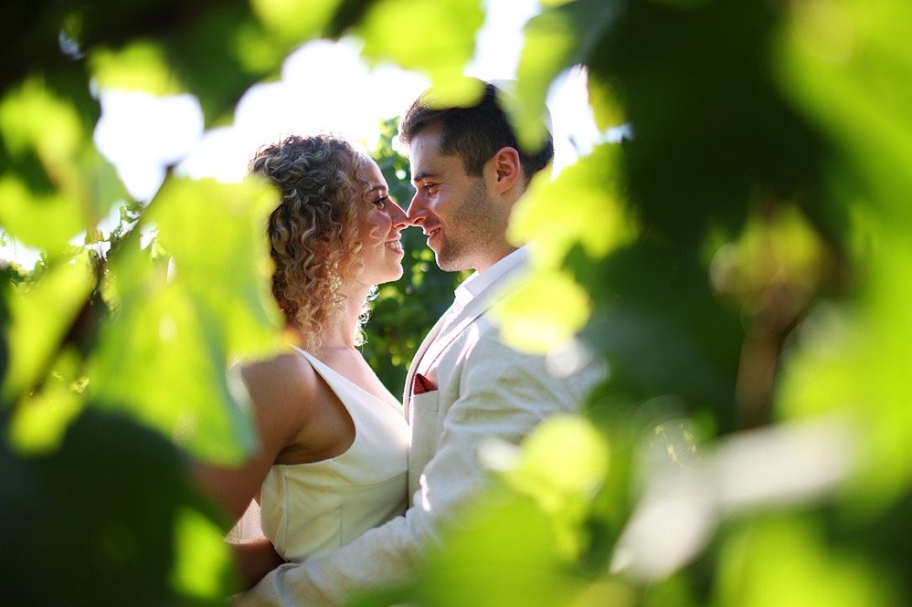 A couple dressed in white, smiling and touching noses, surrounded by green leaves and foliage.