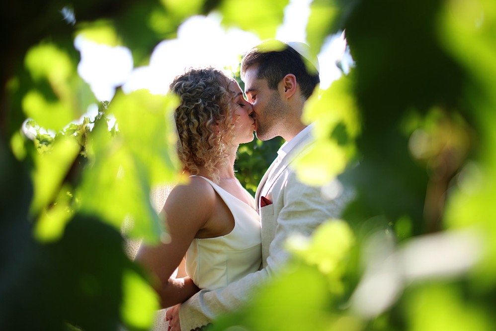 A couple sharing a kiss outdoors surrounded by green leaves.