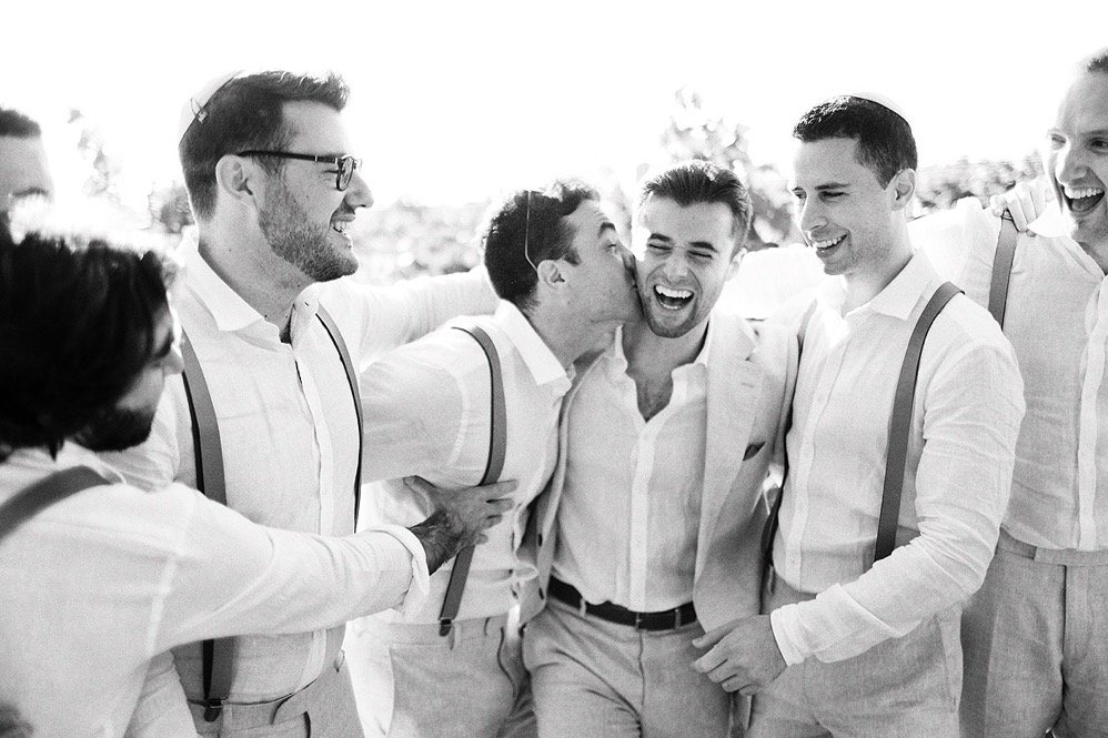 A group of seven men in light-colored clothing, smiling and laughing together outdoors, one man kissing another on the cheek.
