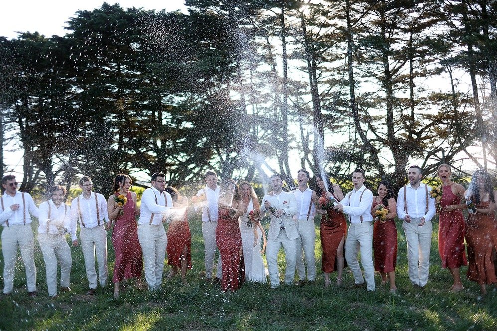 Group of people celebrating outdoors in a wooded area, dressed in formal attire with some holding flowers, as water is sprayed or splashed during the celebration.