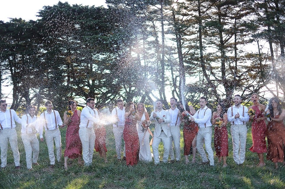 Group of people celebrating outdoors, spraying champagne, with trees and sunlight in the background.