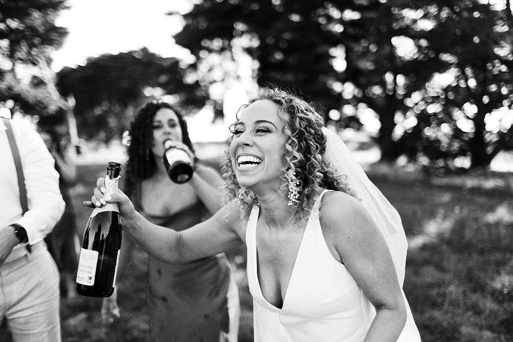 A woman in a wedding dress laughing and holding a champagne bottle outdoors during a celebration, with two other women in the background also enjoying drinks.