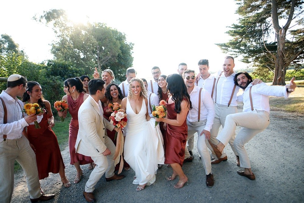 Group of people celebrating outdoors at a wedding, with a bride in a white dress, bridesmaids in maroon dresses, and groomsmen in white shirts with suspenders, holding flowers and smiling.