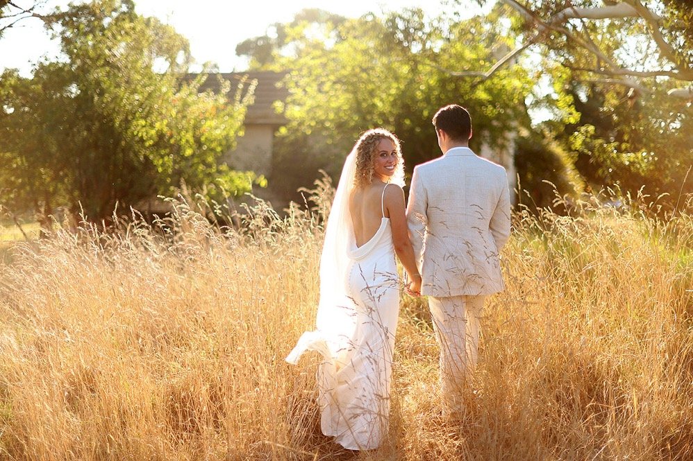 A bride and groom walk hand in hand through a field of tall golden grass, with trees and a house in the background, during a sunny outdoor wedding.