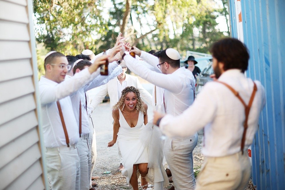 A group of men dressed in white with suspenders and yarmulkes are celebrating outdoors, forming an arch with beer bottles for a woman in a wedding dress to walk through. The woman is smiling joyfully.