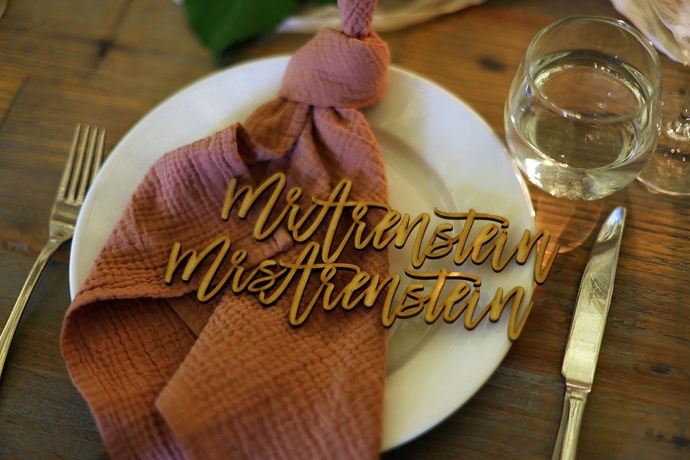 A white plate with a pink cloth napkin tied in a knot, placed on a wooden table. There is a gold-colored word sign that reads 'MIA FRIENDSHIP' on the napkin. The setting includes a fork on the left, a knife on the right, and a glass of water on the t