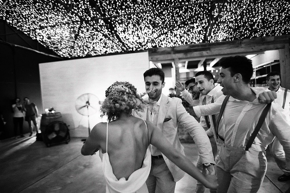 People dancing at a wedding reception, with a woman in a backless dress and floral headpiece, and men in suits and suspenders, under decorative string lights.