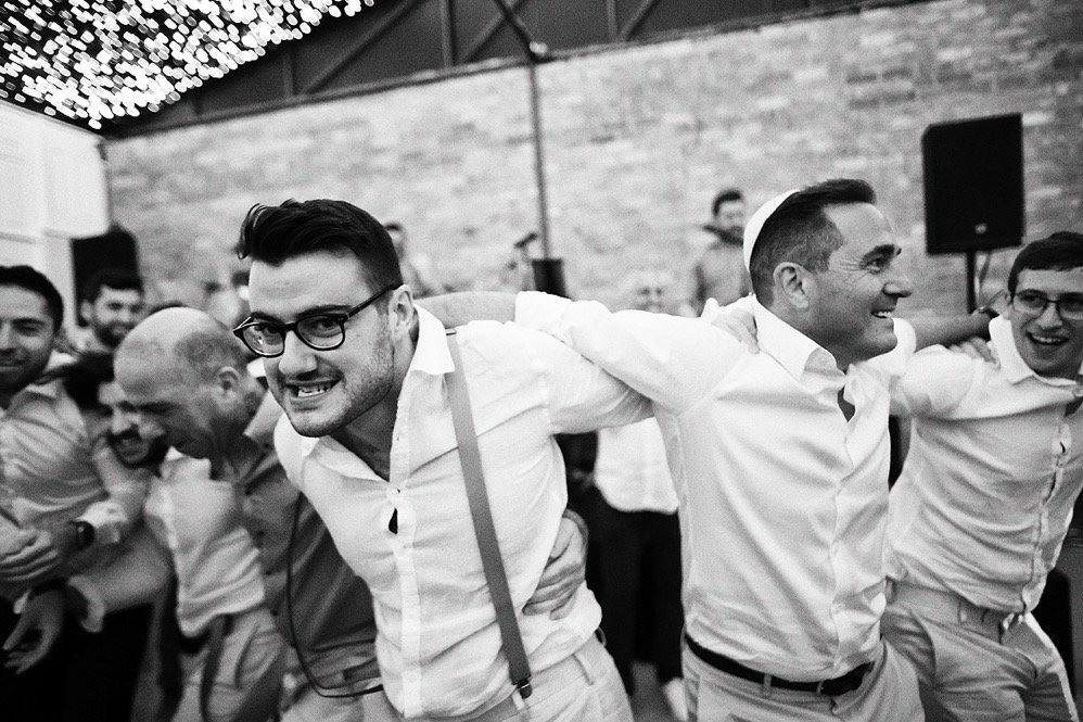 Group of men in white shirts with their arms around each other, smiling and enjoying a celebration or party in a decorated indoor venue.