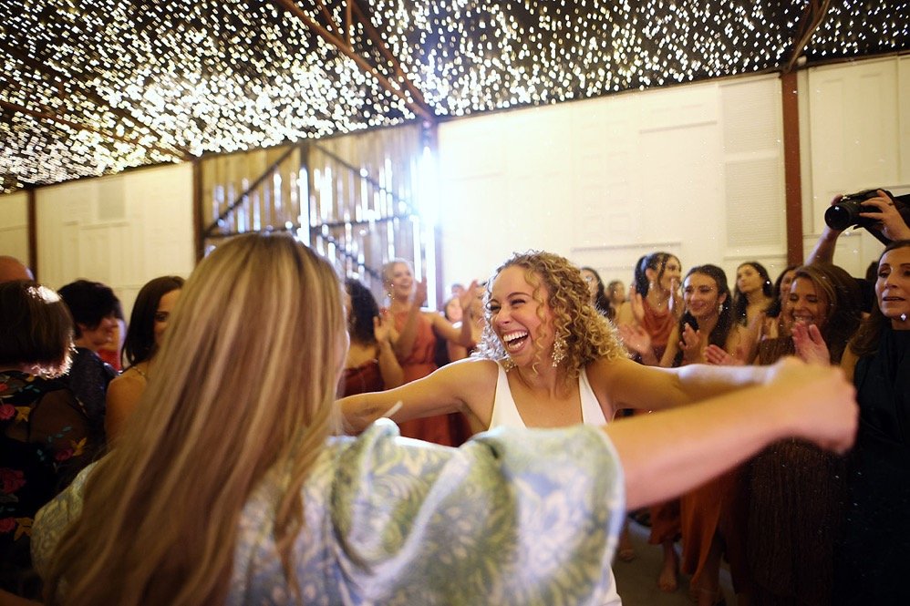 A group of women dancing and laughing at a celebration or party in a decorated barn with string lights.