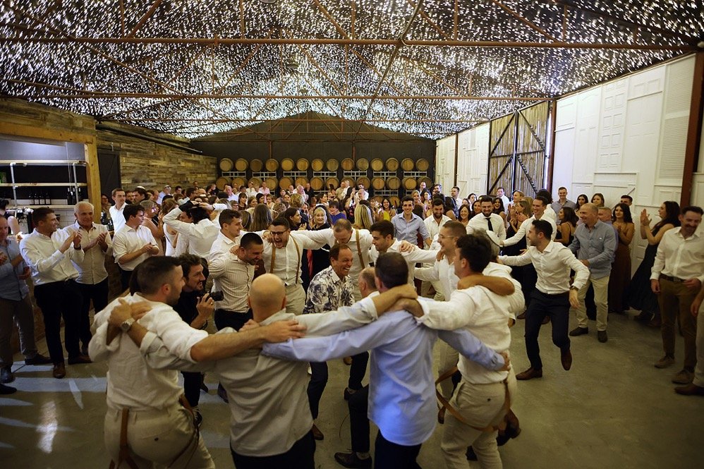 Group of people dancing in a circle at a celebration or party with string lights overhead.