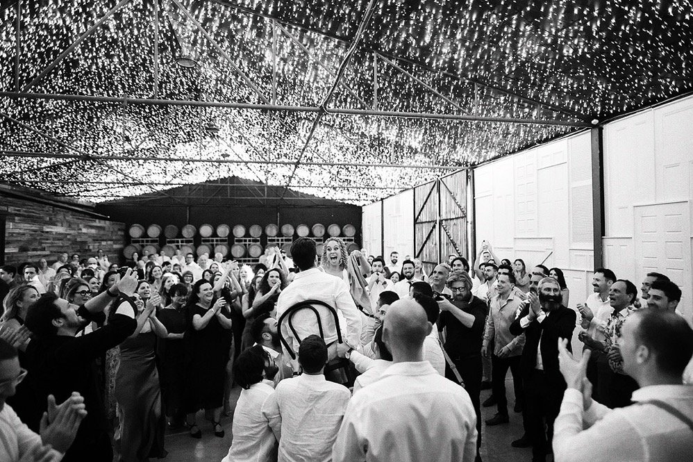 Groom carrying bride on his shoulders during wedding celebration with guests clapping and cheering in a decorated venue with string lights on the ceiling.