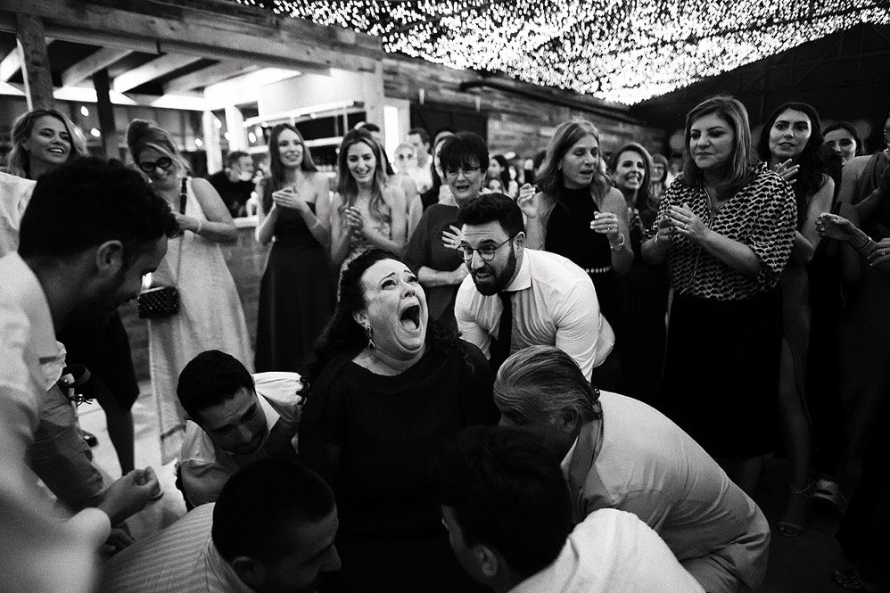 Group of people celebrating at an event, with a woman in the center laughing and surrounded by others with smiles, under a decorated ceiling with string lights.
