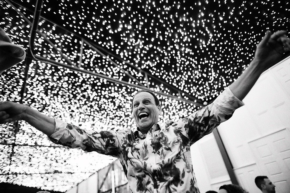 A man with his arms raised and smiling in a festive indoor setting decorated with hanging string lights.