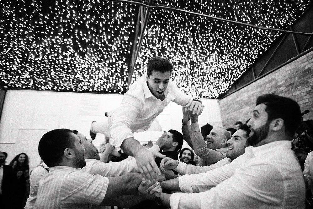 People at a celebration dance together, lifting a man in the air under a ceiling decorated with string lights.