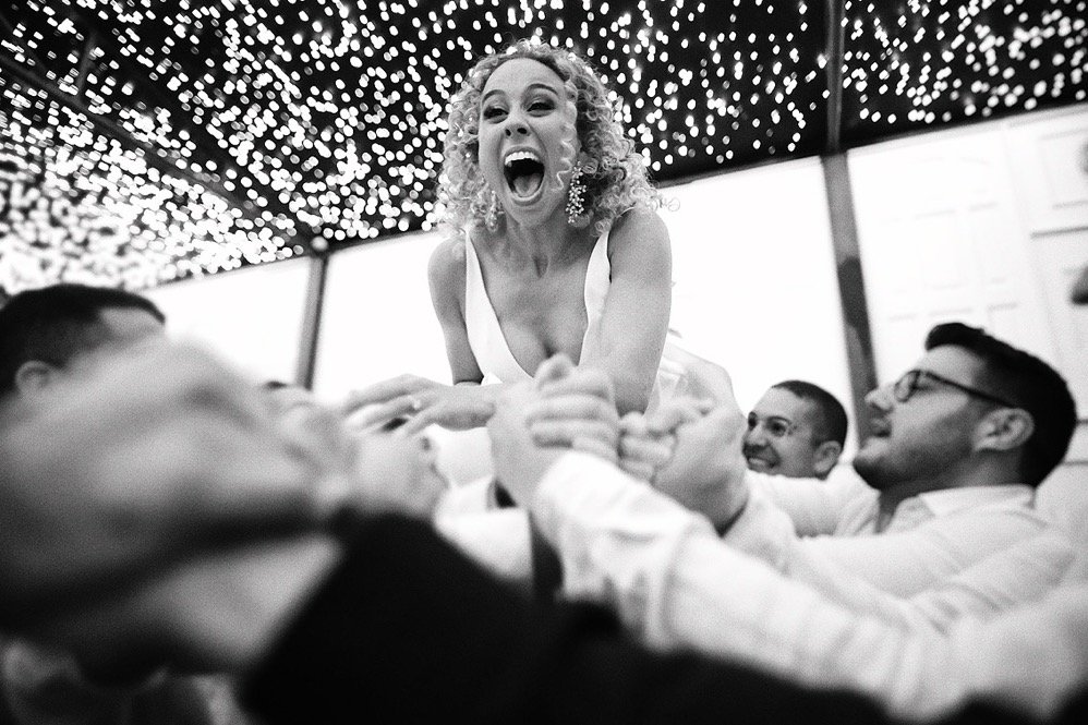 A woman with curly hair is smiling and laughing while being lifted by a group of men at a celebration or party, with string lights overhead.