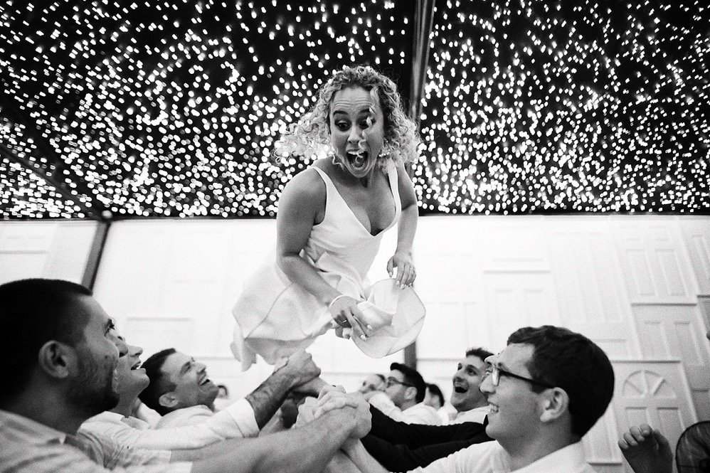 Women in a white dress being lifted by a group of men in a celebration setting with twinkling lights overhead.