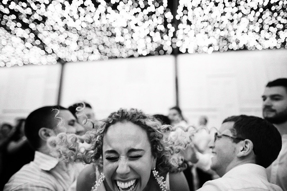A woman with curly hair laughing joyfully at a crowded party or celebration under a decorated ceiling with shimmering lights.