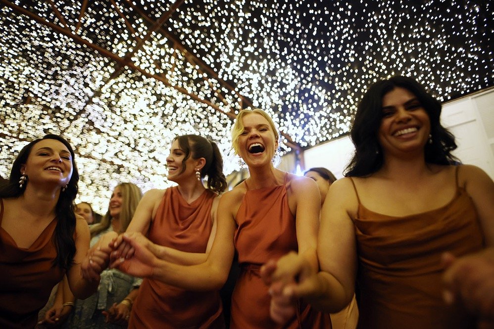 A group of women in matching brown dresses smiling and holding hands at an event with a ceiling decorated with numerous small string lights.