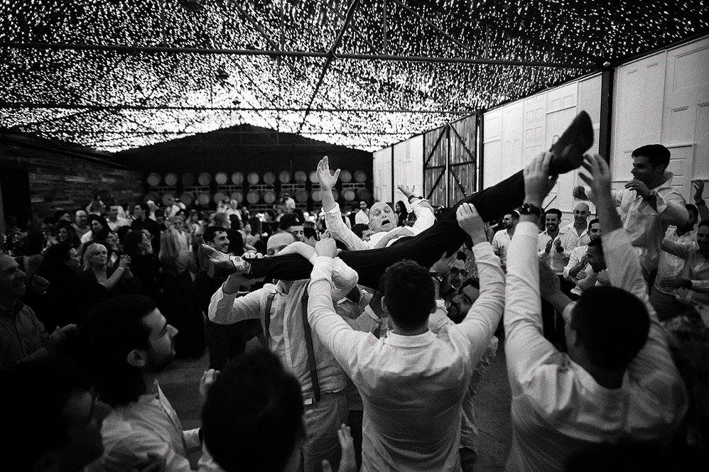 A lively celebration with multiple guests lifting a woman horizontally in a barn decorated with hanging lights.