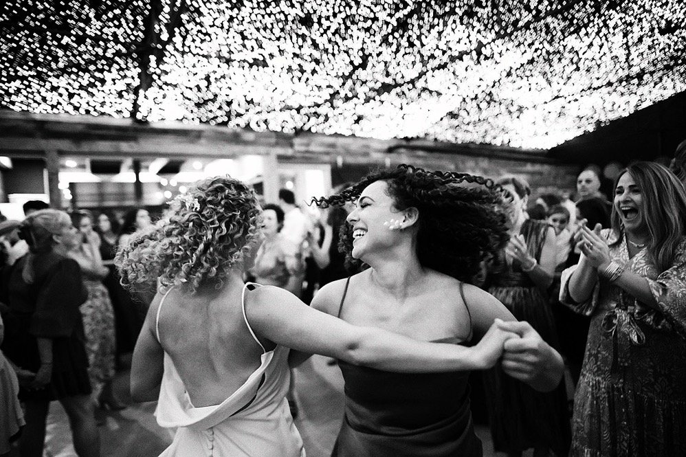 Two women dancing and smiling at a party, surrounded by other guests, with string lights overhead.