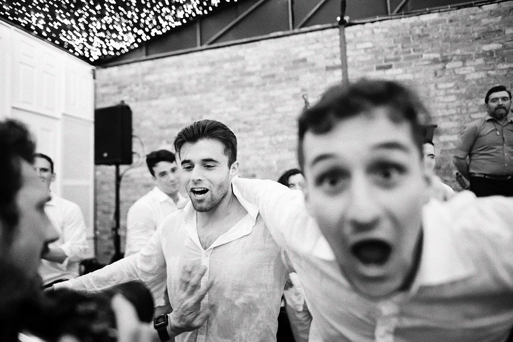 A group of young men in white shirts having fun at a social gathering or party, with some dancing and enjoying themselves in a venue with brick walls and string lights.