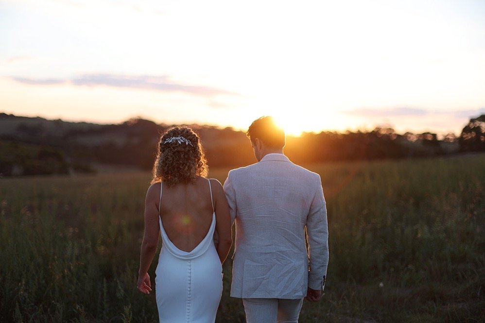 A couple walking through a field during sunset, with the sun setting behind them.