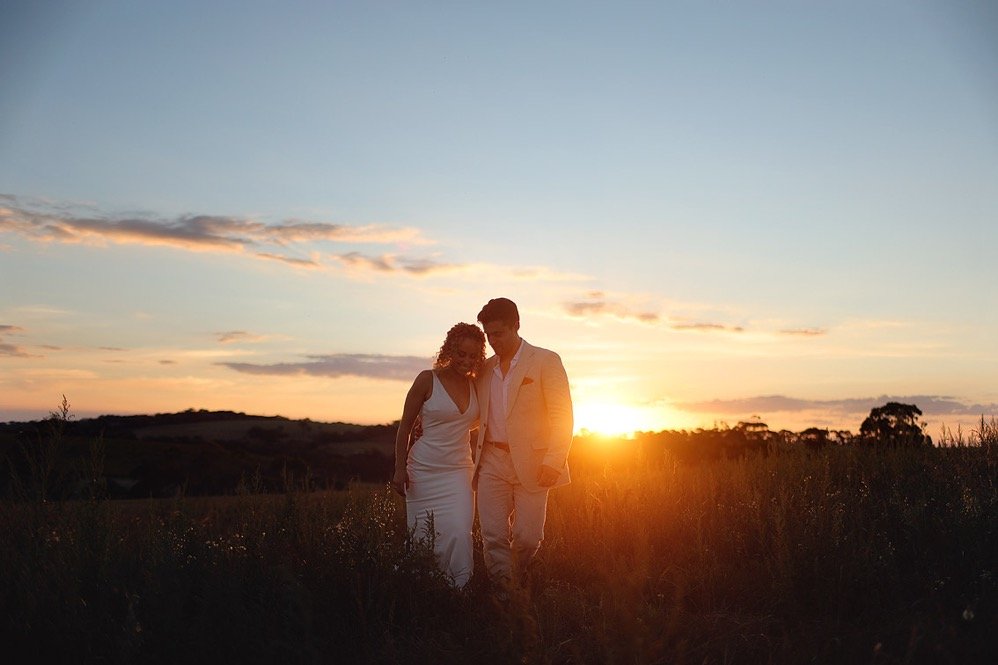 A couple walking together in a field at sunset, with the sun low on the horizon and a partly cloudy sky.