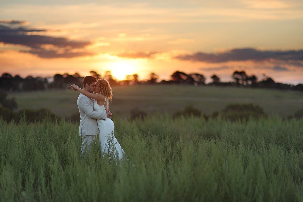 A couple embracing in a field during sunset, with trees and a colorful sky in the background.