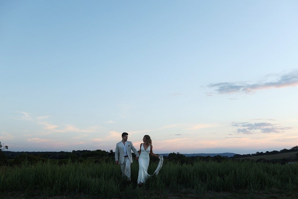 A couple in wedding attire walking hand-in-hand through a grassy field during sunset.
