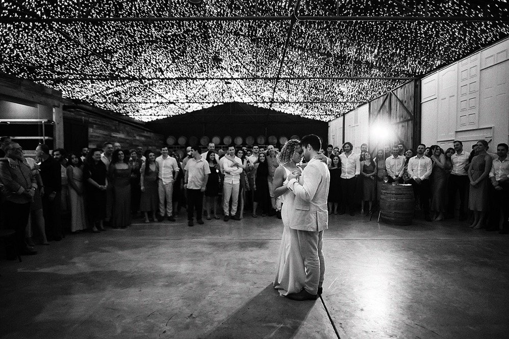 A bride and groom dancing closely in the center of a wedding reception, with guests standing in a circle around them under string lights display.