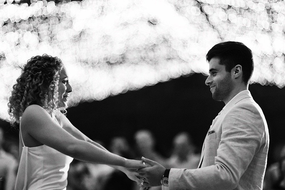 A black-and-white photo of a couple holding hands and smiling at each other during a dance or celebration, with blurred string lights in the background.