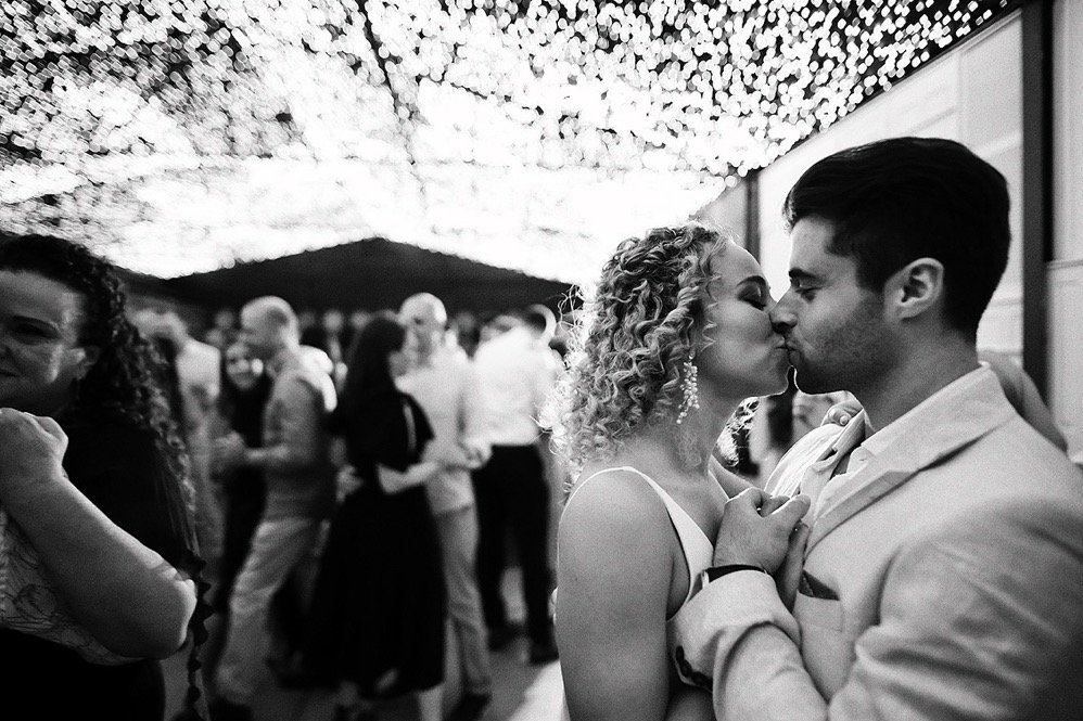 A couple is kissing at a wedding reception under hanging string lights, with guests in the background.