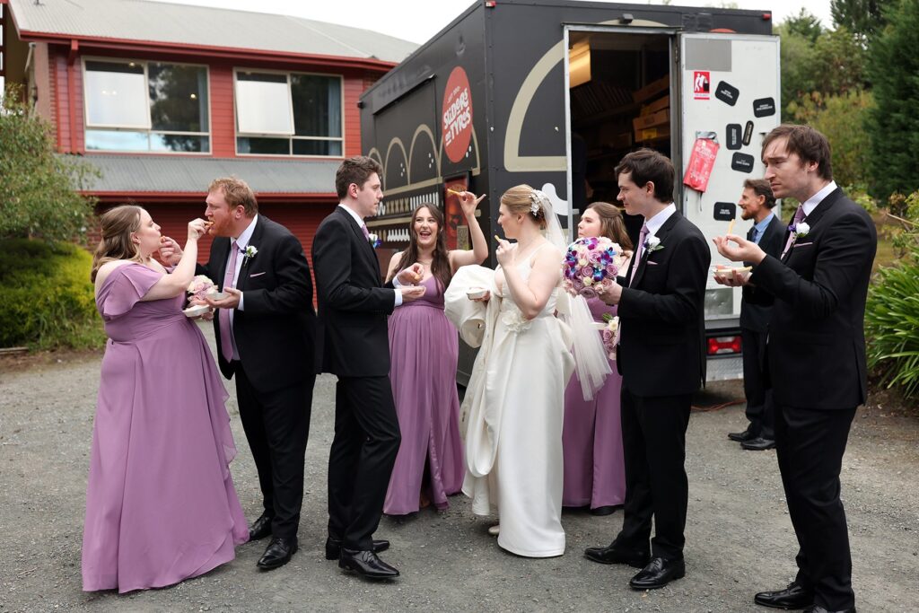 Bride, groom and attendants eat food beside a black catering truck outside a building.