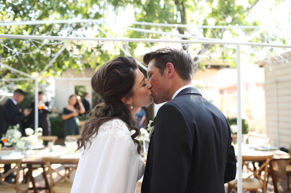 Bride in a white lace dress and groom in a dark suit kiss at an outdoor reception.